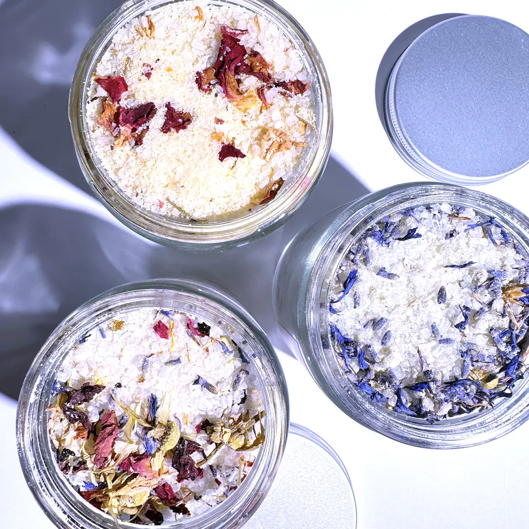 Three glass jars of By Valenti Goat's Milk Bath Salts with dried flower petals on a light background