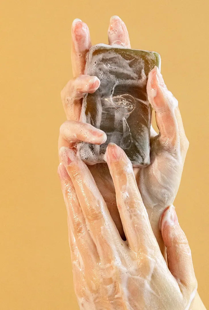 A woman's hands holding a By Valenti Japanese Matcha Bar Soap covered in foam in front of a yellow background
